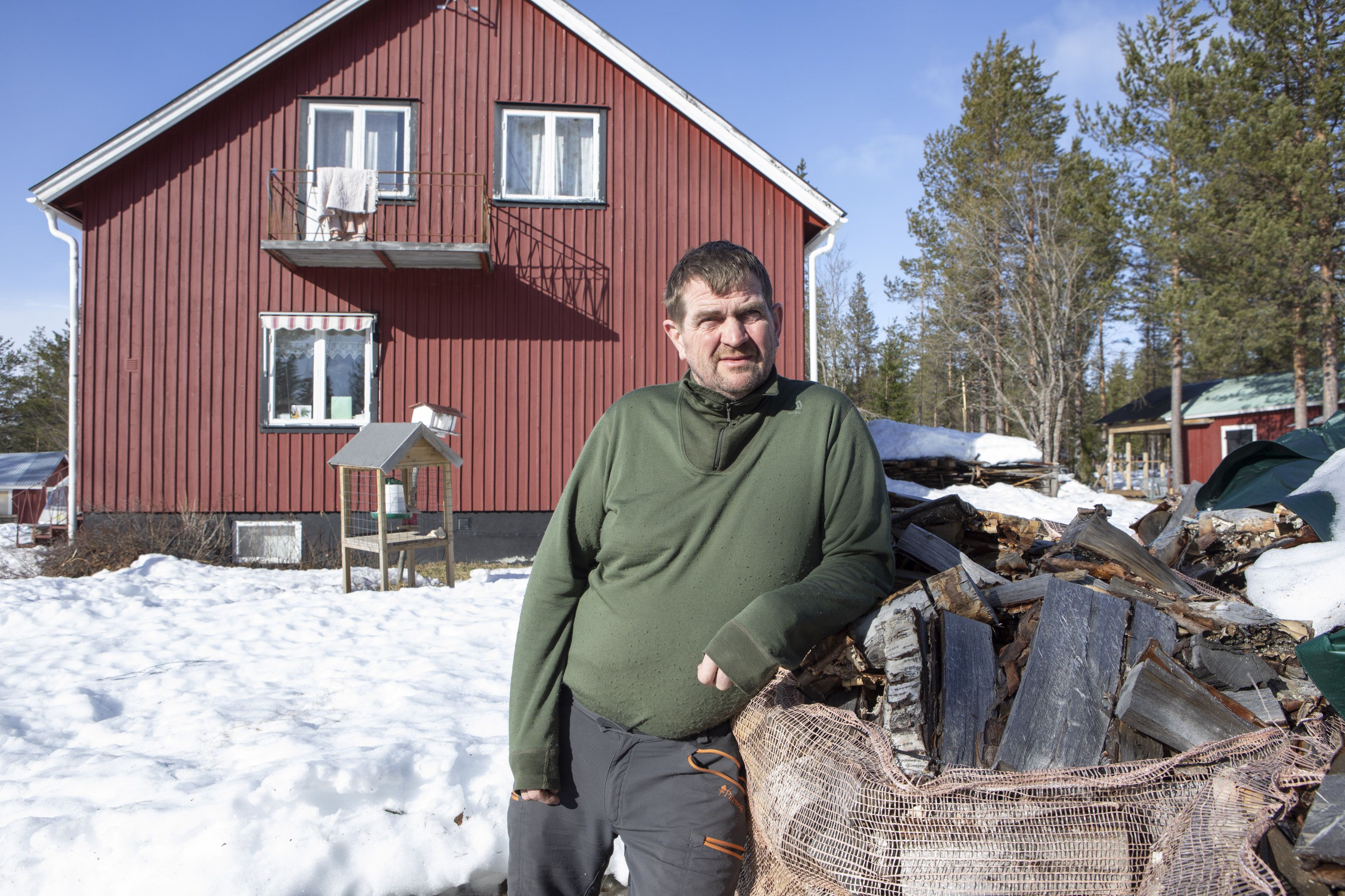 Man lutar sig mot vedhög framför rött hus i snöigt landskap