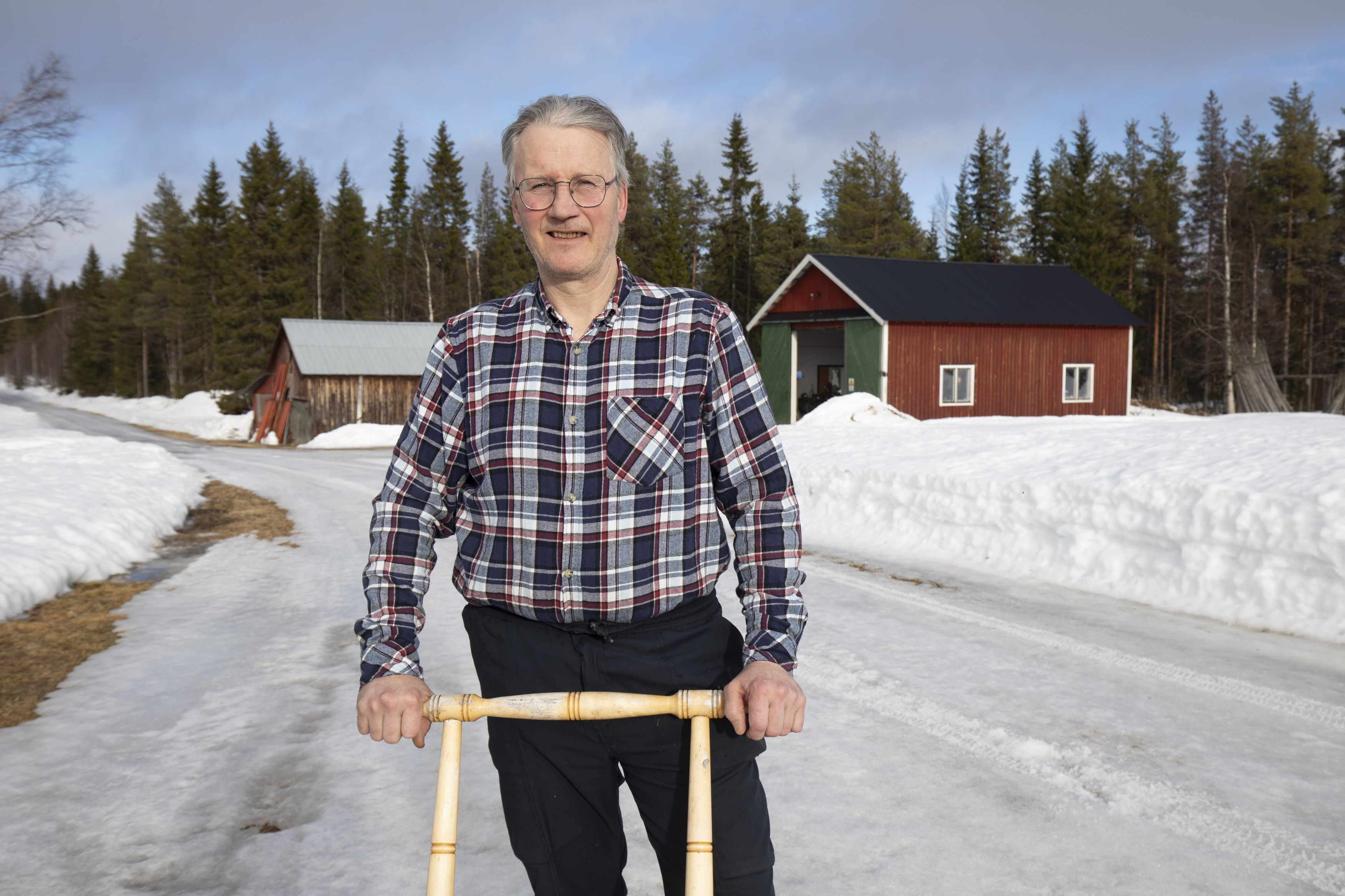 Äldre man står ute på snöig landsbygdsväg med spark framför röda lador
